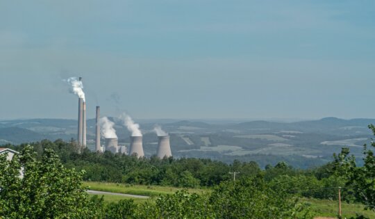 Johnstown, PA, USA - June 12, 2008: Mixed coal and nuclear power plant in Appalachian wilderness under light blue sky. Chimneys and cool towers spew white vapor clouds. Green foliage up front.